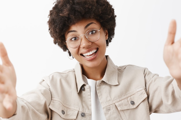 Kostenloses Foto nahaufnahmeaufnahme der freundlichen emotionalen und niedlichen afroamerikanerfrau in den transparenten gläsern und im hemd, die hände in richtung ziehen, um selfie zu nehmen, kopf zu neigen, lächelnd, das breites neues foto online postend