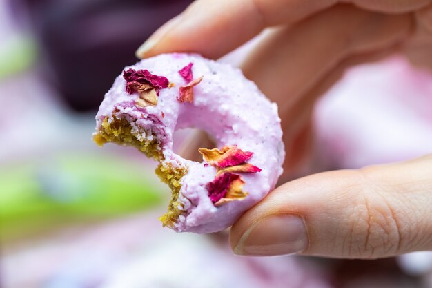 Nahaufnahmeaufnahme der Hand einer Frau, die einen kleinen veganen Donut hält, der mit essbaren Rosenblumen bedeckt ist