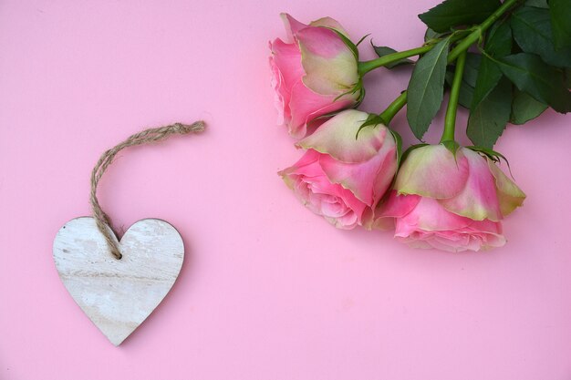 Nahaufnahmeaufnahme der rosa Rosenblumen mit einem Herzholzanhänger mit Raum für Text auf einer rosa Oberfläche