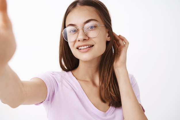 Nahaufnahmeaufnahme der selbstbewussten gut aussehenden sinnlichen und weiblichen jungen Frau, die Haar hinter Ohr flirty lächelt, freudig tragend Brille tragend Selfie genießt Blick der schönen sauberen Haut