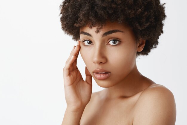 Nahaufnahmeaufnahme der sinnlichen und zarten schönen Afroamerikanerfrau mit reiner Haut, die Gesicht sanft berührt und mit niedlichen und liebevollen Emotionen nackt posiert