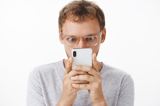 Nahaufnahmeaufnahme des amüsierten und unterhaltenen männlichen Geeks in der Brille, die Smartphone nah an Gesicht knallenden Augen hält und fasziniert auf Gerätebildschirm starrt, der schockierende interessante Nachricht über weißer Wand liest