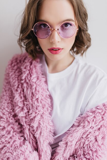 Nahaufnahmeaufnahme des brünetten kurzhaarigen Mädchens in der lila Sonnenbrille, die im Pelzmantel aufwirft. Innenporträt des sinnlichen Mädchens in der rosa flauschigen Jacke lokalisiert auf weißer Wand.