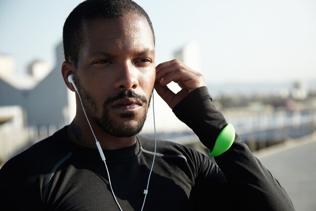 Nahaufnahmeaufnahme des jungen schwarzen Mannes mit Bart, der Kopfhörer in sein Ohr setzt. Entschlossener Sportler ist bereit für Langstreckenlauf und Training bei Sonnenaufgang. Athlet, der Sport-Fitness-Tracker trägt.