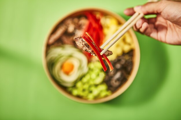 Nahaufnahmeaufnahme einer menschlichen Hand, die ein Stück Fleisch mit rotem Pfeffer durch chinesische Stöcke hält