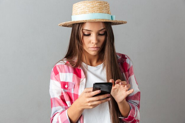 Nahaufnahmefoto der ernsten jungen Frau im Strohhut, Telefon betrachtend,