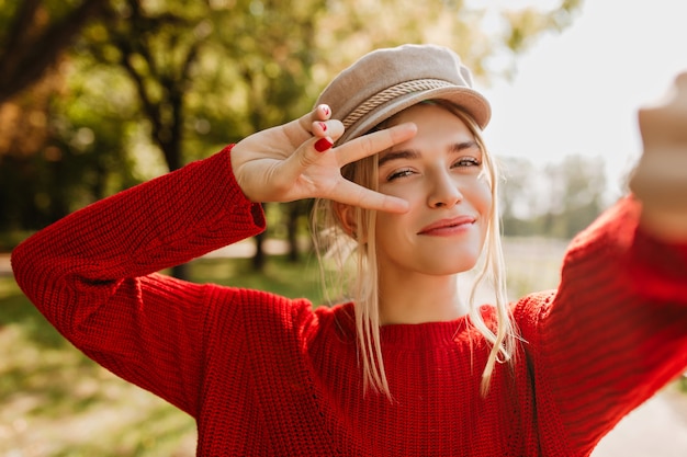 Nahaufnahmefoto der glänzenden schönen Blondine im trendigen roten Pullover und im hellen Hut, die glückliches selfie im Herbst machen.