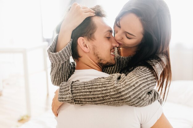 Nahaufnahmefoto der romantischen Dame in der grauen Kleidung, die Freund auf hellem Hintergrund umarmt