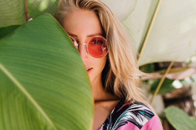 Nahaufnahmefoto der sinnlichen blonden Frau in der rosa Sonnenbrille, die sich hinter grünem Blatt versteckt.