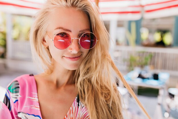 Nahaufnahmefoto des freudig gebräunten weiblichen Modells mit langen blonden Haaren. Porträt der begeisterten blonden Dame in der rosa Sonnenbrille, die auf unscharfem Hintergrund aufwirft.