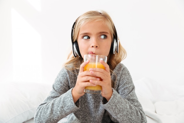 Nahaufnahmefoto des hübschen kleinen Mädchens im Kopfhörer lecken, während es Orangensaft trinkt, beiseite schauend