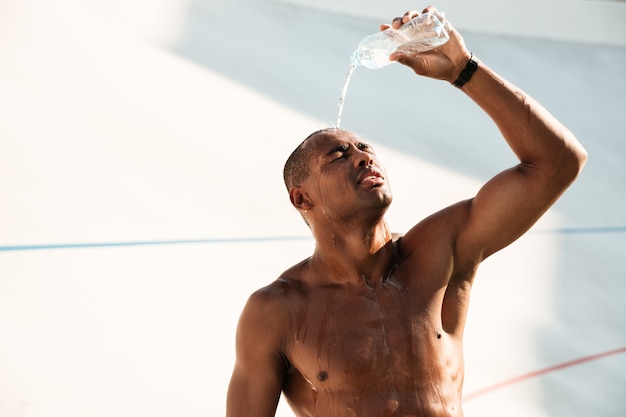 Nahaufnahmefoto des jungen afrikanischen sportmannes, der wasser auf seinem kopf ausschüttet und nach dem training ruht