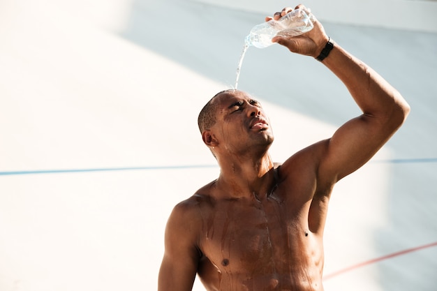 Nahaufnahmefoto des jungen afrikanischen Sportmannes, der Wasser auf seinem Kopf ausschüttet und nach dem Training ruht