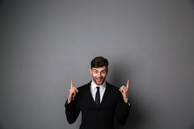 Nahaufnahmefoto des jungen lächelnden Mannes in der formellen Abnutzung, die mit zwei Fingern nach oben zeigt