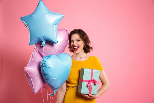 Nahaufnahmefoto des verspielten Geburtstagskindes mit den roten Lippen zwinkert ein Auge, während Geschenkbox und Luftballons halten