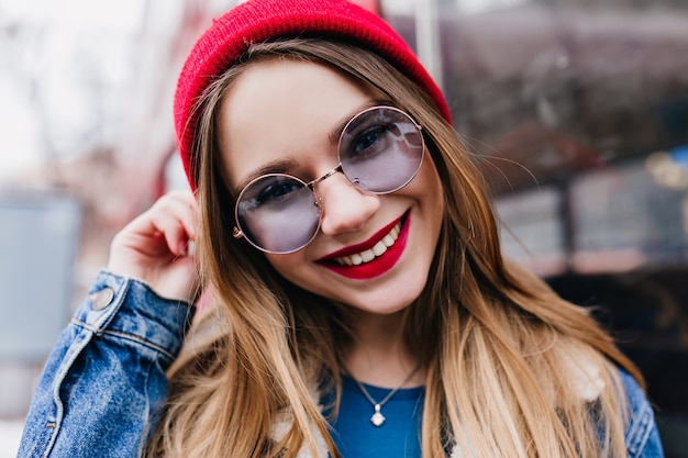 Nahaufnahmefoto des weißen Mädchens, das in der Stadt im Frühlingswochenende entspannt. Außenaufnahme der wunderbaren europäischen Dame in der Jeansjacke und in der blauen Brille.