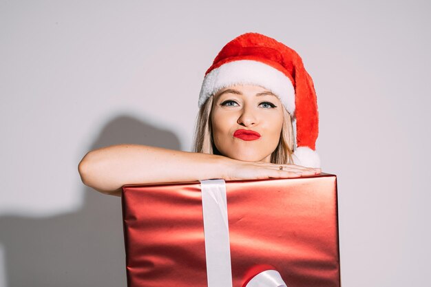 Nahaufnahmefoto einer jungen blonden dame, die sankt-hut und roten lippenstift trägt, während sie sich auf ein weihnachtsgeschenk stützt. urlaubskonzept