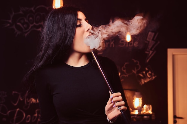 Nahaufnahmefoto eines verführerischen brünetten Mädchens im schwarzen Oberteil, das eine Wasserpfeife in einem Nachtclub oder einer Wasserpfeifenbar raucht.