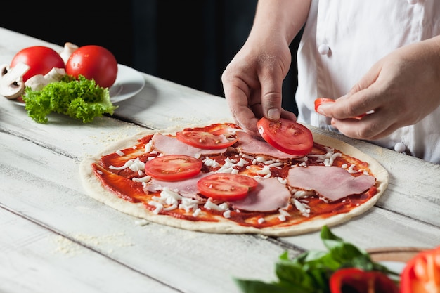 Nahaufnahmehand des Kochbäckers in der weißen Uniform, die Pizza an der Küche macht