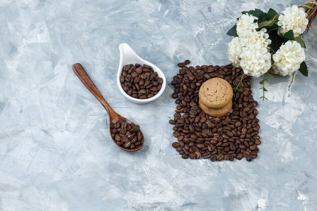 Nahaufnahmekaffeebohnen im hölzernen Löffel, weißer Porzellankrug mit Keksen, Blumen auf hellblauem Marmorhintergrund. horizontal