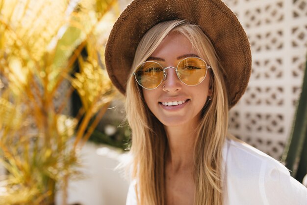 Nahaufnahmeporträt der angenehmen weißen Frau trägt eleganten Sommerhut. Außenaufnahme der positiven blonden Frau in den trendigen gelben Gläsern, die Urlaub genießen.