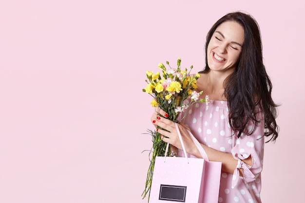 Nahaufnahmeporträt der attraktiven dunkelhaarigen jungen Frau im Kleid, genießt den Frühling, mag Blumen und Geschenke von ihrem hasband bekommen, Augen glücklich schrauben. Menschen, Geschenke, Feierkonzept.