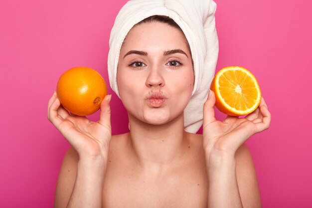 Nahaufnahmeporträt der attraktiven fröhlichen Frau hält orange Scheiben, hält Lippen gefaltet, trägt Handtuch und nackte Schultern, posiert auf rosa. Model posiert im Studio. Natürliches Schönheitskonzept.