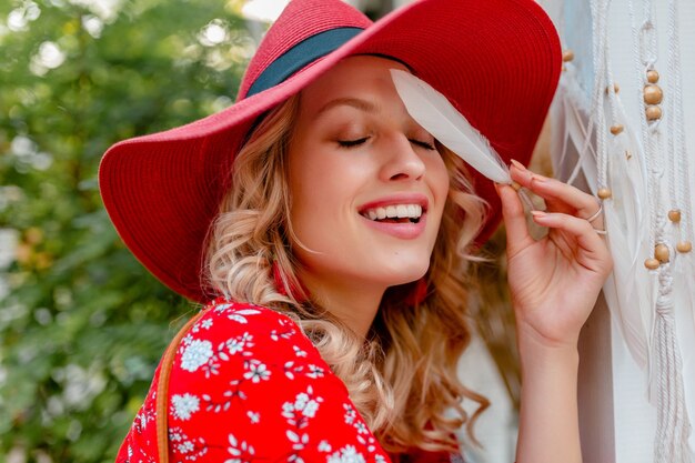 Nahaufnahmeporträt der attraktiven stilvollen blonden lächelnden Frau im strohroten Hut und im Blusensommer-Mode-Outfit, das sexy sinnliche Gesichtshaut der weißen Feder hält