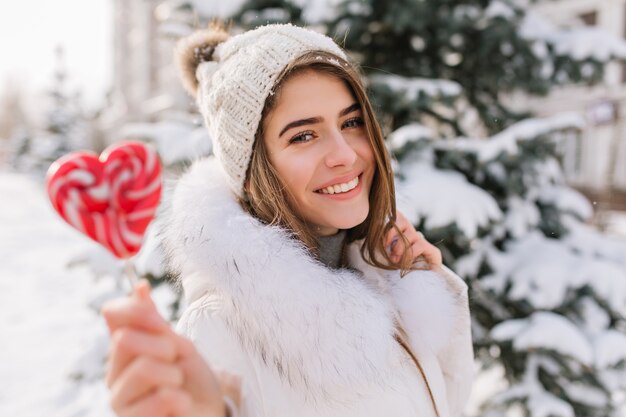 Nahaufnahmeporträt der bezaubernden Dame im weißen Mantel, der süßen Lutscher hält. Foto im Freien der glückseligen blonden Frau in der Strickmütze, die neben Baum am Wintertag mit roter Kandiszucker aufwirft.