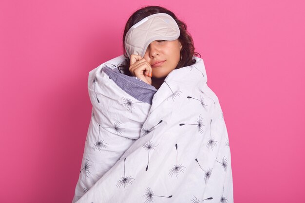 Nahaufnahmeporträt der brünetten Frau, die von der Schlafmaske guckt, nicht aufwachen will, Augen geschlossen hält und weiße Decke trägt