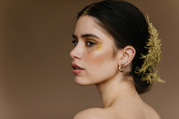 Nahaufnahmeporträt der charmanten frau trägt goldene ohrringe. süßes brünettes mädchen mit pflanze im haar.