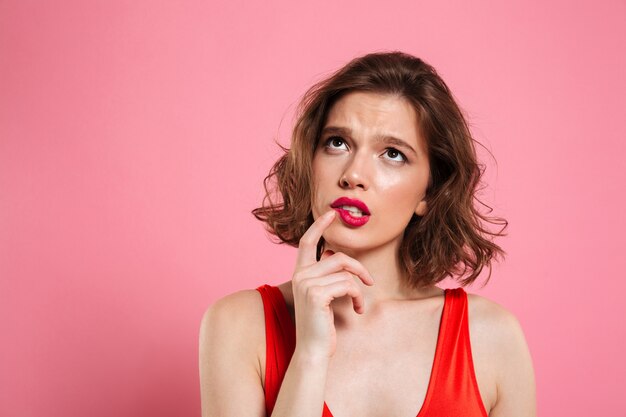 Nahaufnahmeporträt der denkenden jungen schönen Frau mit den roten Lippen, die mit dem Finger ihre Wange berühren und nach oben schauen