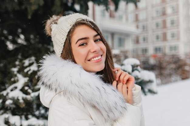 Nahaufnahmeporträt der erstaunlichen Frau in der weißen Strickmütze mit Schneeflocken auf den Haaren lächelnd auf der Straße.