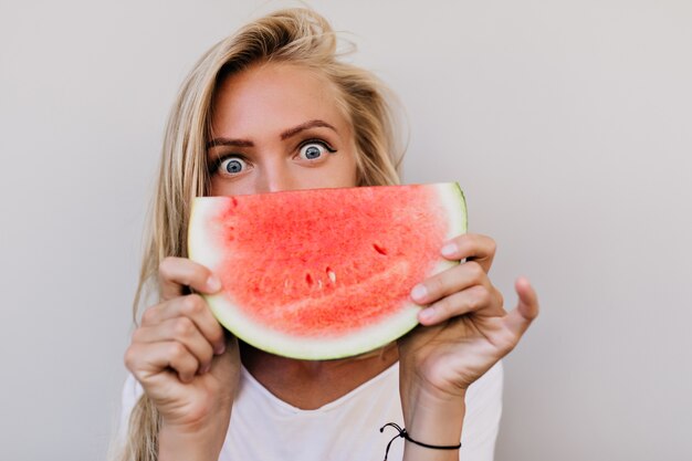 Nahaufnahmeporträt der Frau mit den grauen Augen, die Wassermelone essen. Innenfoto der spektakulären kaukasischen Dame mit langer Frisur, die Früchte genießt.