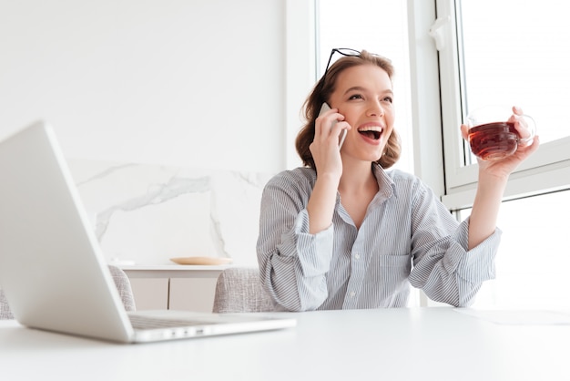 Nahaufnahmeporträt der glücklichen brünetten Frau im gestreiften Hemd, das auf Handy spricht, während Tasse des Tees drinnen hält