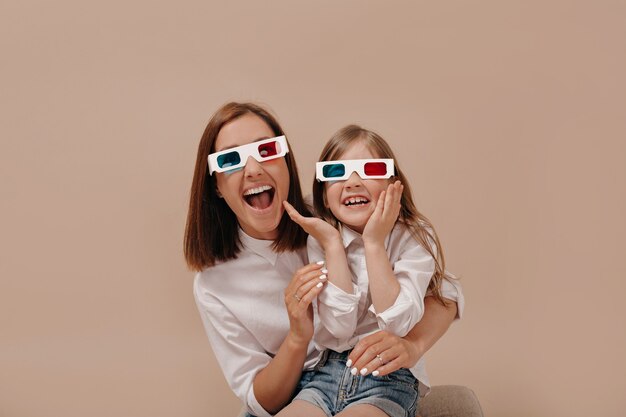 Nahaufnahmeporträt der glücklichen Frau mit dem kleinen Mädchen, das einen Film in der 3D-Brille mit überraschten Emotionen ansieht