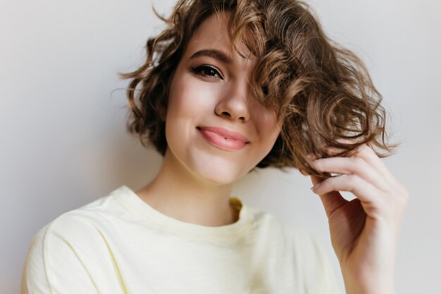 Nahaufnahmeporträt der glücklichen jungen Frau mit dem trendigen Make-up, das mit den kurzen Haaren spielt. Innenfoto des bezaubernden lockigen Mädchens lokalisiert auf weißer Wand.