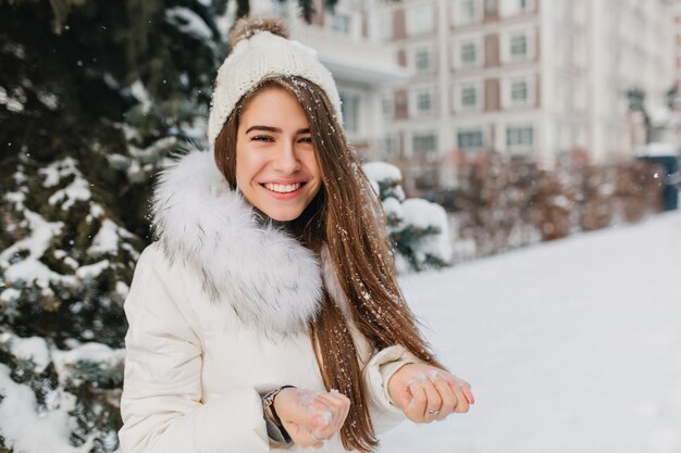 Nahaufnahmeporträt der herrlichen blonden Frau, die Schnee in den Händen hält und lächelt. Spektakuläre Frau, die Wintermorgen im Hof genießt und mit jemandem spielt.