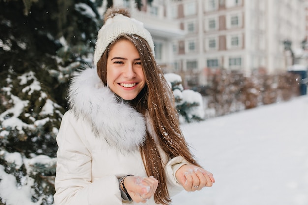 Nahaufnahmeporträt der herrlichen blonden Frau, die Schnee in den Händen hält und lächelt. Spektakuläre Frau, die Wintermorgen im Hof genießt und mit jemandem spielt.