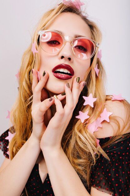 Nahaufnahmeporträt der herrlichen Blondine, die auf der Party sexy aussieht, bedeckt mit Sternkonfetti, sinnlich berührendes Gesicht. Trägt eine rosa trendige Brille, hat langes lockiges Haar, rote Lippen und eine stilvolle Maniküre. Isoliert..