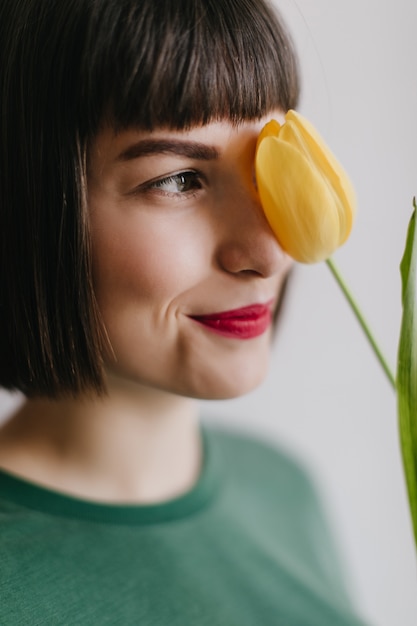 Nahaufnahmeporträt der herrlichen brünetten Dame mit hellem Make-up, das mit gelber Blume aufwirft. fröhliche weiße Frau mit Tulpe.