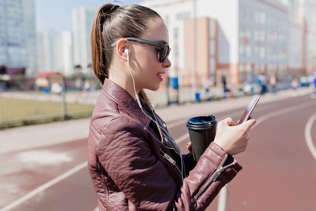 Nahaufnahmeporträt der hübschen Dame mit dem dunklen gesammelten Haar trägt schwarze Sonnenbrille, trinkt Kaffee, schaut in das Telefon und hört Musik in Kopfhörern