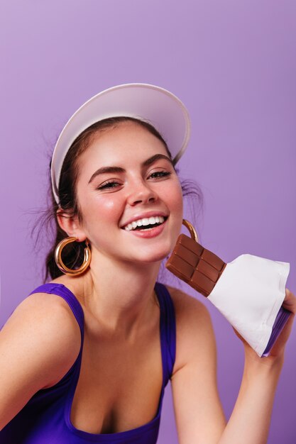 Nahaufnahmeporträt der jungen Frau in den hellen Goldohrringen und in der weißen Kappe lächelnd und hält Tafel Schokolade auf lila Wand
