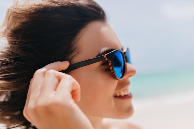 Nahaufnahmeporträt der jungen Frau in der Sonnenbrille, die auf Unschärfennatur aufwirft. Faszinierende kaukasische Frau mit dunklem Haar, das Sommer im Seebad genießt.