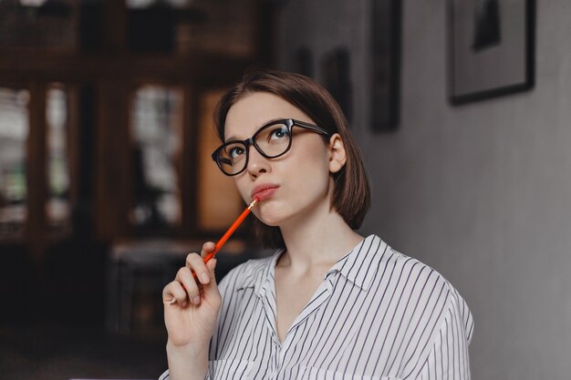 Nahaufnahmeporträt der jungen kurzhaarigen Geschäftsfrau in den Gläsern, die nachdenklich nach oben schauen und roten Stift halten.