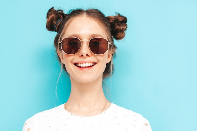 Nahaufnahmeporträt der jungen schönen lächelnden Frau im trendigen grünen Sommerkleid Sexy sorglose Frau mit der Frisur mit zwei Hörnern, die in der Nähe der blauen Wand im Studio posiert Positives Modell mit Sonnenbrille