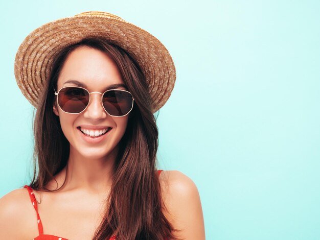 Nahaufnahmeporträt der jungen schönen lächelnden Frau im trendigen SommerkleidSexy sorglose Frau posiert in der Nähe der blauen Wand im StudioPositives Modell, das sich drinnen amüsiertFröhlich und glücklich mit Sonnenbrille und Hut