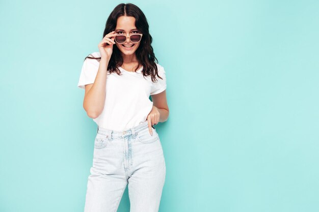 Nahaufnahmeporträt der jungen schönen lächelnden Frau in der modischen Sommerkleidung Sexy sorglose Frau, die nahe der blauen Wand im Studio aufwirft Positives Modell, das Spaß drinnen hat Sonnenbrille ausziehen