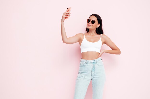 Nahaufnahmeporträt der jungen schönen lächelnden Frau in der modischen Sommerkleidung Sexy sorglose Frau, die nahe rosa Wand im Studio aufwirft Positives Modell, das Spaß drinnen hat Selfie-Fotos mit Sonnenbrille machen