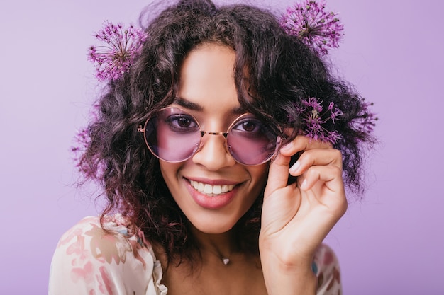 Nahaufnahmeporträt der lächelnden braunäugigen Frau mit Blumen im schwarzen Haar. Afrikanische glückselige Dame in der Sonnenbrille, die aufwirft.